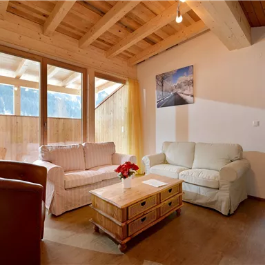 A cozy living room with a wooden ceiling and large windows. There are comfortable sofas and a wooden table with a floral arrangement.