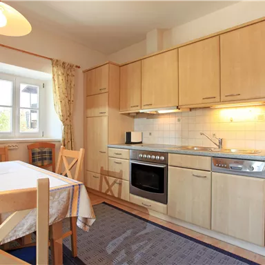 Eine helle Küche mit Holzmöbeln und moderner Ausstattung. Am Tisch steht ein Tischgedeck und das Fenster lässt Tageslicht herein.