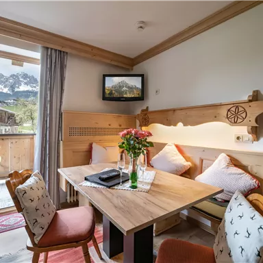 Ein gemütlicher Wohnraum mit einem Holztisch und Stühlen. Durch die offene Balkontür sieht man einen Blick auf die Berge.