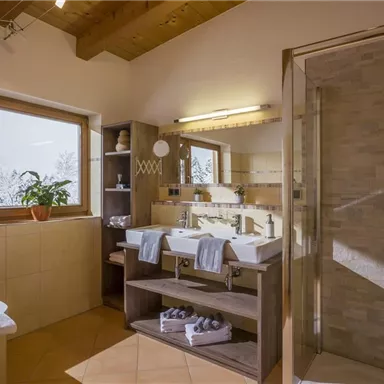 Ein modernes Badezimmer mit zwei Waschbecken und einer großen Dusche. Helle Fliesen und ein Fenster bringen viel Licht herein.