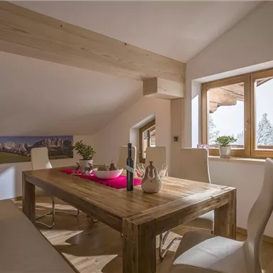 Ein gemütlicher Essbereich mit einem großen Holztisch und weißen Stühlen. Durch das Fenster sieht man eine verschneite Landschaft.