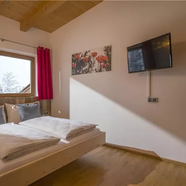 Ein gemütliches Schlafzimmer mit einem großen Bett und zwei Fenstern. An der Wand hängt ein Fernseher und ein farbenfrohes Bild.