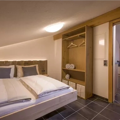 Ein gemütliches Schlafzimmer mit einem Doppelbett, Holzdetails und moderner Einrichtung. Der Raum ist gut beleuchtet und hat einen Zugang zu einem weiteren Bereich.