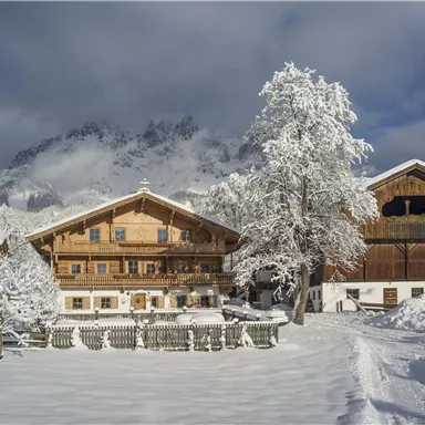 Eine malerische Winterlandschaft mit schneebedeckten Hütten und Bäumen. Im Hintergrund erheben sich majestätische Berge unter einem klaren Himmel.