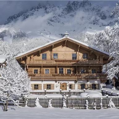 Ein charmantes Holzhaus im Schnee, umgeben von verschneiten Bäumen. Im Hintergrund sind majestätische Berge zu sehen.