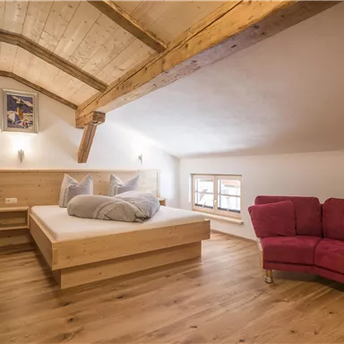 Ein gemütliches Schlafzimmer mit Holzdecke und einem großen Bett. Ein rotes Sofa und Fenster sorgen für eine helle Atmosphäre.