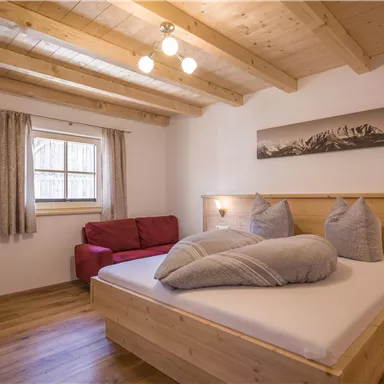 Ein gemütliches Schlafzimmer mit Holzdecke und einem Doppelbett. Ein rotes Sofa und ein Fenster sorgen für eine helle Atmosphäre.