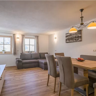 Ein modernes Wohnzimmer mit einer gemütlichen Couch und einem Esstisch. Helle Fenster sorgen für eine freundliche Atmosphäre.