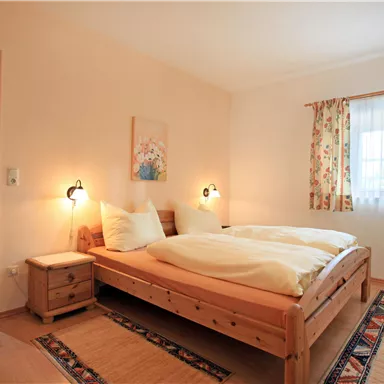 Ein gemütliches Schlafzimmer mit einem Doppelbett und zwei Nachttischen. Sanfte Farben und eine helle Fensterdekoration sorgen für eine einladende Atmosphäre.