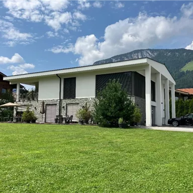Ein modernes Haus mit großen Fenstern und einem gepflegten Garten. Im Hintergrund sind grüne Berge und ein blauer Himmel sichtbar.