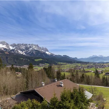 Eine malerische Landschaft mit schneebedeckten Bergen und grünen Wiesen. Im Vordergrund ist ein schönes Haus und Bäume zu sehen.