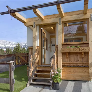 Ein charmantes Holzhaus mit einer einladenden Eingangstür und einer Veranda. Im Hintergrund sind Berge und ein klarer Himmel zu sehen.