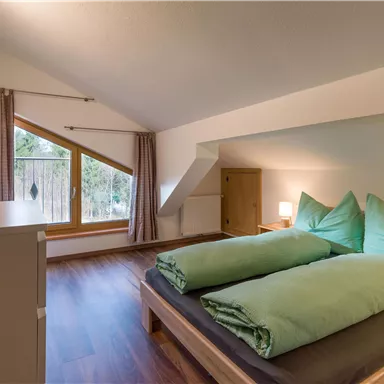 Ein gemütliches Schlafzimmer mit zwei Betten und hellgrünem Bettzeug. Große Fenster lassen viel Tageslicht herein und bieten einen Blick ins Grüne.