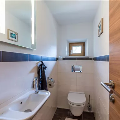 Ein modernes Badezimmer mit einer Toilette, einem Handwaschbecken und einem Spiegel. Die Wände sind hell und es gibt ein kleines Fenster für natürliches Licht.