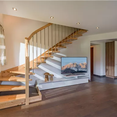 Ein modernes Wohnzimmer mit einer Holztreppe und einem großen Fernseher. Die Wände sind hell gestrichen und es gibt natürliches Licht durch die Fenster.