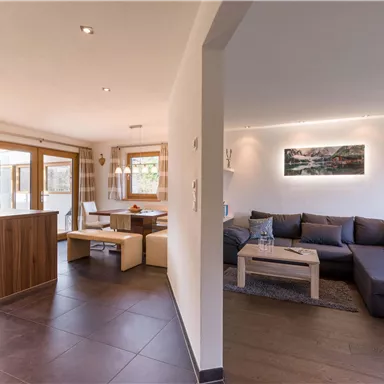 Ein modernes Wohnzimmer mit einem gemütlichen Sofa und einem Tisch. Der offene Raum bietet einen Blick auf die Küche und hat große Fenster, die Tageslicht hineinlassen.