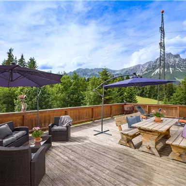 Eine gemütliche Terrasse mit Sitzgelegenheiten und Sonnenschirmen. Im Hintergrund sind Wälder und Berge zu sehen, unter einem blauen Himmel.