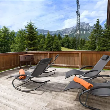 Ein gemütlicher Balkon mit Liegestühlen und orangefarbenen Handtüchern. Im Hintergrund sind hohe Bäume und die Berge zu sehen.