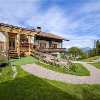 Ein rustikales Haus inmitten der Natur mit grünem Rasen. Im Hintergrund sind Berge und ein klarer Himmel zu sehen.