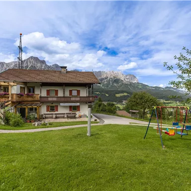Ein gemütliches Haus in einer grünen Landschaft mit einem Spielplatz. Im Hintergrund sind majestätische Berge und ein blauer Himmel zu sehen.