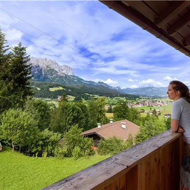 Eine Frau steht auf einem Balkon und schaut auf die schöne Berglandschaft. Im Hintergrund sind grüne Wiesen und bewaldete Hügel zu sehen.