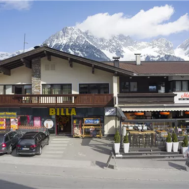 Ein Geschäftshaus in den Bergen mit dem Logo "BILLA" im Erdgeschoss. Im Vordergrund stehen parkende Autos und eine Terrasse mit Tischen und Stühlen.