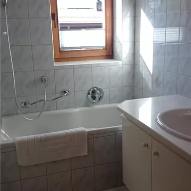 Ein helles Badezimmer mit einer Badewanne, einem Waschbecken und einem Fenster. Die Fliesen sind in hellen Farben gehalten und sorgen für eine freundliche Atmosphäre.