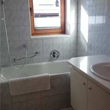 Ein helles Badezimmer mit einer Badewanne, einem Waschbecken und einem Fenster. Die Fliesen sind in hellen Farben gehalten und sorgen für eine freundliche Atmosphäre.