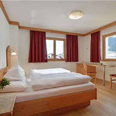 A cozy hotel room with wooden furniture and bright walls. There is a large window with a view of the mountains and a small seating area.