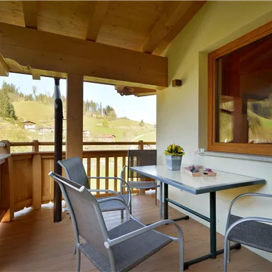 A cozy balcony with a table and two chairs. In the background, green meadows and trees can be seen.