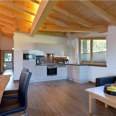 A modern kitchen with bright wooden ceilings and large windows. A wooden dining table stands next to a cozy seating area.