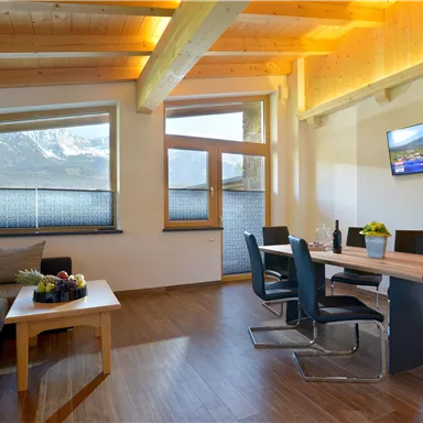 A modern living room with a wooden ceiling and large windows. There is a sofa, a dining table, and a television on the wall.