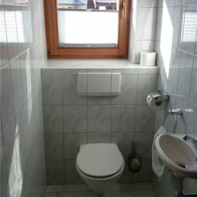 Ein kleines Badezimmer mit weißen Fliesen und einem Fenster. Es gibt eine Toilette und ein Waschbecken.
