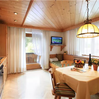 Eine gemütliche Küche mit Holzvertäfelung und einem Tisch, der mit Obst und einer Flasche Wein gedeckt ist. Ein kleiner Fernseher hängt an der Wand und helle Vorhänge lassen viel Licht herein.