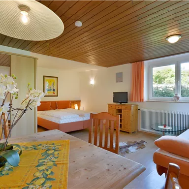 Ein gemütliches Zimmer mit einem Essbereich, einem Sofa und einem Doppelbett. Große Fenster lassen viel Tageslicht herein.