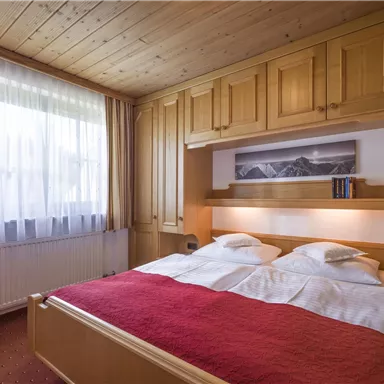 Ein gemütliches Schlafzimmer mit einem Doppelbett und Holzschränken. Große Fenster lassen viel Tageslicht herein.