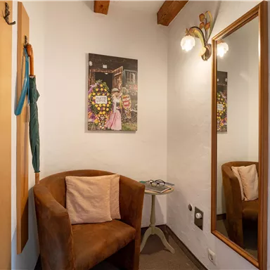 Ein gemütlicher Eingangsbereich mit einem braunen Sessel und einem großen Spiegel. An der Wand hängt ein Bild einer Frau mit Blumen.