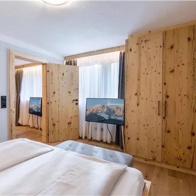 Ein modernes Schlafzimmer mit einem Holzkleiderschrank und zwei Fernsehern. Die Fenster sind mit Vorhängen dekoriert und lassen viel Tageslicht herein.