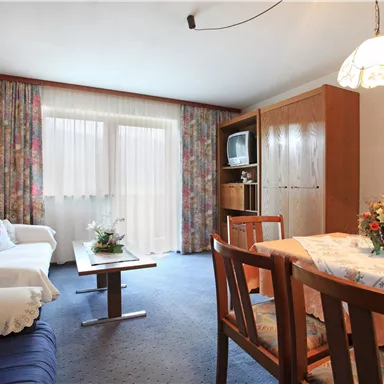 A cozy living room with a blue sofa and a dining table. Bright curtains let in plenty of light and create an inviting atmosphere.