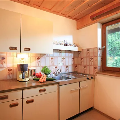 A cozy kitchen with wooden paneling and a window that looks out onto nature. Fresh vegetables and kitchen utensils are on the countertop.