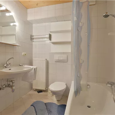 Ein modernes Badezimmer mit einer Badewanne, einem Waschbecken und einem Spiegel. Die Wände sind hell gefliest und es gibt einen blauen Badezimmerteppich.