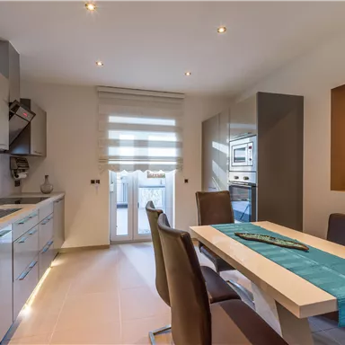 Eine moderne Küche mit einer hellen Farbpalette und eleganten Möbeln. Der Tisch ist mit einer blauen Tischdecke dekoriert und es gibt viel natürliches Licht.