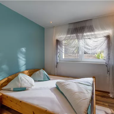 Ein helles Schlafzimmer mit einem Doppelbett und Holzlattenboden. Die Wände sind in einem beruhigenden Blau gestrichen und ein Fenster lässt viel Tageslicht herein.