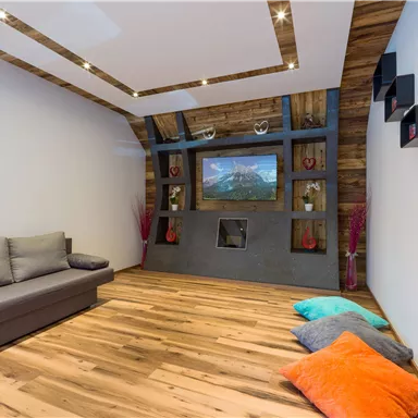 Ein modernes Wohnzimmer mit einem grauen Sofa und einem großen Fernseher an einer Holzvertäfelung. Bunte Kissen auf dem Boden verleihen dem Raum eine einladende Atmosphäre.