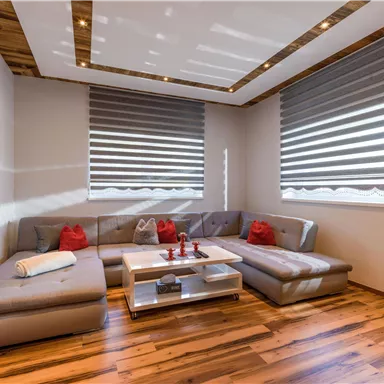 Ein modernes Wohnzimmer mit einem großen, gemütlichen Ecksofa und roten Kissen. Der Holzboden und die eleganten Jalousien sorgen für eine warme Atmosphäre.