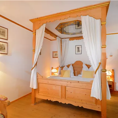 A cozy bedroom with a four-piece wooden bed and white curtains. The room has warm lighting and traditional decorative elements.