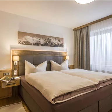 A cozy room with a large double bed and stylish wooden decor. Large windows provide plenty of daylight and a view of the mountains.