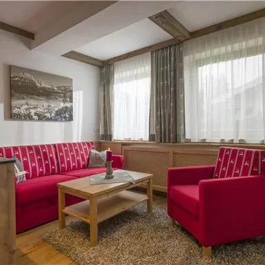 A cozy living room with red sofas and a wooden table. Large windows provide plenty of daylight and an inviting atmosphere.