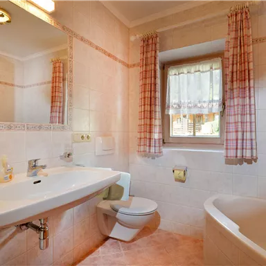 Ein helles Badezimmer mit einem Waschbecken, einer Toilette und einer Badewanne. Die Wände sind gefliest und es gibt Vorhänge an den Fenstern.