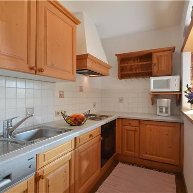 Eine moderne Küche mit Holzschränken und einer hellen Arbeitsfläche. Auf der Theke steht eine Obstschale, und der Raum hat ein Fenster mit Vorhängen.